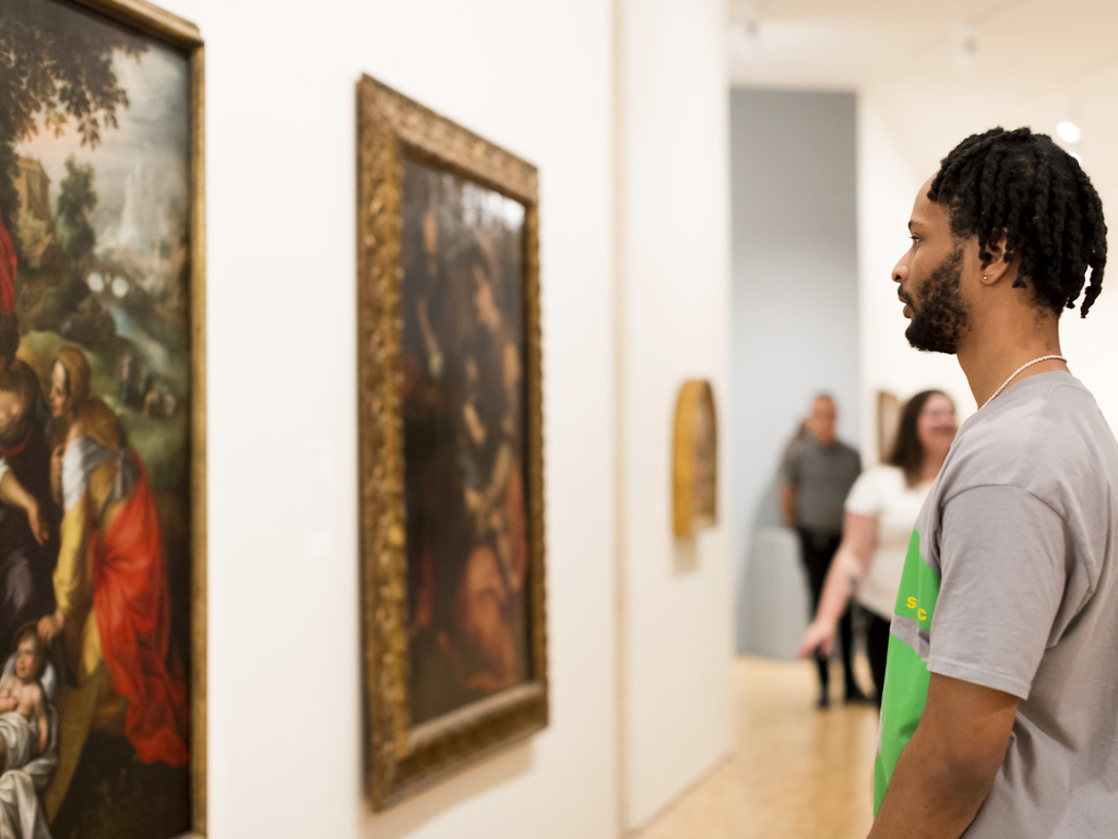 A young man stands in front of a painting hanging on the wall.  He is looking at the painting.