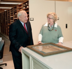 A man in a suit with a red tie and a woman in a green and white blouse with colorful jewelry stand together next to a tall table. On the table in front of them is a sepia-colored print in a wooden frame.