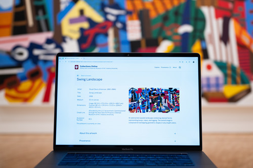 A laptop sits in front of a large, out of focus mural in this photo. The mural has intertwined tones of blue, red, white, and black. The laptop is open to a webpage of the mural's object record on the Eskenazi Museum's website. 
